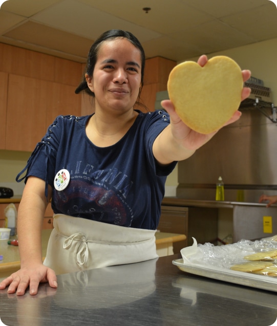 A woman in a kitchen holds out a heart cookie to the camera, smiling.